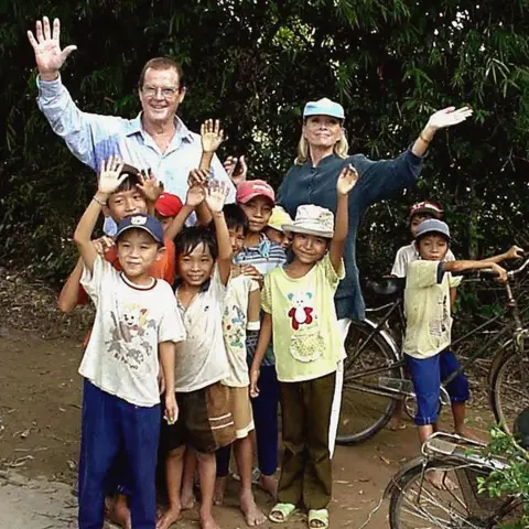 Getty Images Sir Roger Moore and his fourth wife Kristina are posing for a picture with 10 children in Vietnam. They are all waving at the camera and smiling. Sir Roger is 75 and is wearing glasses and a checked shirt. His wife Kristina is wearing a grey jumper and has a blue baseball cap on her head. One child is sitting astride a bicycle, while the others, all dressed in t-shirts and shorts or trousers, are standing in front of Sir Roger and Kristina looking happy.