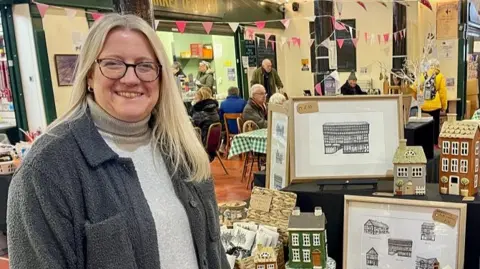 A lady stands by a market stall covered in paintings, drawings and model clay houses. She has long blonde hair, glasses, a grey fleece jacket and a cream jumper. Behind her, people are sat eating and drinking in the market cafe with bunting hanging above their heads.