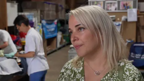 Svetlana Ryzhevska, in a green top and with white blonde hair, looking to the side of the camera in a warehouse.. Behind her volunteers are looking through donations. 