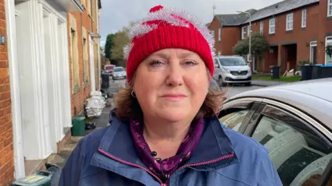 Ben Schofield/BBC Jane Carr, looking direct to camera and standing on the pavement of a residential street. Jane is wearing a red and silver festive bobble hat and a blue waterproof coat, with a purple coloured top underneath. She has blue eyes and her light brown hair can be seen emerging from under the red hat. Immediately to the right of her in the image is the side of a car. A silver van is parked further away behind her on the opposite side of the road. To the left of the image are the frontages of terraced houses. On the pavement are some small foodwaste bins, as well as a pile of sandbags.