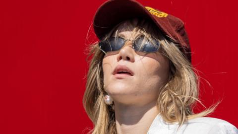 Suki Waterhouse performing on stage in 2023. Suki is a 32-year-old woman with long blonde hair worn loose under a burgundy baseball cap. She wears small dark sunglasses, a white shirt and pearl earrings. She's pictured against a red background. 