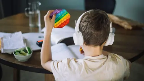 A boy sits at a table with his back to the camera. He is wearing white headphones and there are text books on the table. He has a multicoloured toy in his hand.