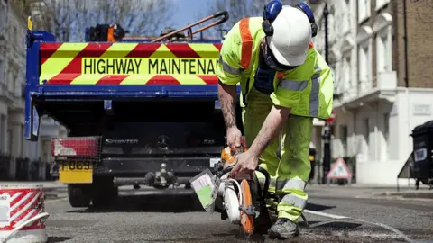 A workman uses a power tool, seemingly a saw, on the road surface. He is leaning forward towards the camera and his face hidden. He is wearing high-viz clothing and a white helmet. Behind him is a trailer, facing away from the camera, which says "Highway Maintenance" on the back.