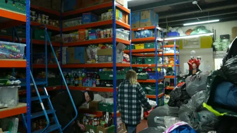 Volunteers organise stock amid rows of shelves stacked with groceries, and bin bags filled with sleeping bags and winter clothes