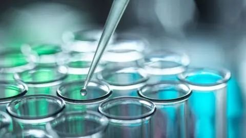 A close-up shot showing the tops of 16 test tubes, set out in a 4x4 square, standing upright. The top of a pipette is in shot, dropping a clear liquid into one of the tubes. There is a blurred blue and green light source behind the tubes, maybe a screen.