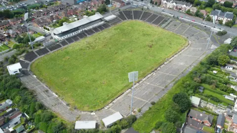 A grass sports pitch with surrounding grey seats and large floodlights. Around the pitch are residential housing.