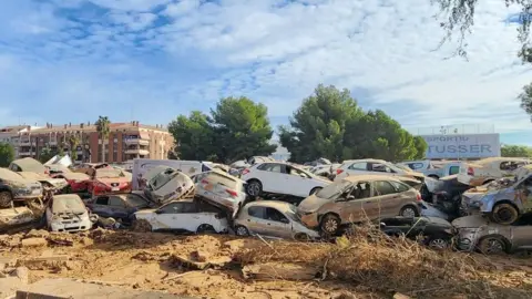 A pile up of around 30 cars which have been left behind in the wake of the flash floods. The ground is muddy and the cars have been smashed and are covered in debris.