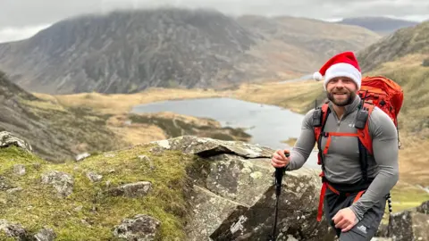 David Glyn Jones Mr Jones pictured smiling wearing a santas hat in the mountains of Eryri National Park. He carries a walking pole in his right hand and has a large backpack on his back. 