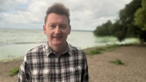 Neil Reid, who has spiky brown hair and a checked shirt stands in front of a green lough