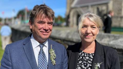 Kirrie and Philip Jenkins smile looking at the camera outside the Royal Chapel in St John's. He is wearing a blue blazer with a white shirt and blue tie with a crisscross pattern in white and a sprig of bollan bane in his button hole. She is wearing a black jacket over a black top with a white floral pattern on it and also has bollan bane pinned to her lapel.