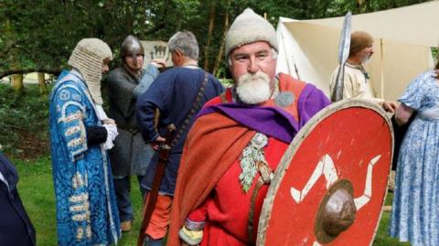 A man dress as a Viking holds a red shield with with a triskelion. Others in traditional dress, including a man in a wig, stand around behind him.