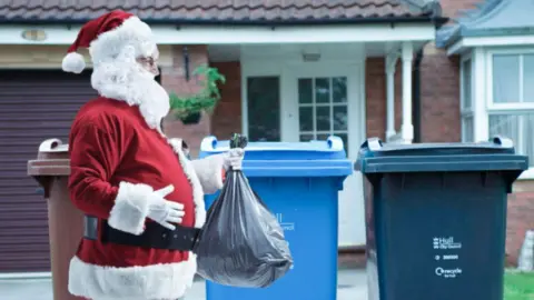 The image shows a person dressed as Santa holding a bin bag with one hand and their belly with the other. They are walking past three bins, one brown, one blue and one black. Behind the bins is a house.