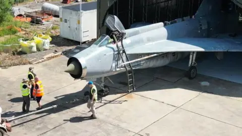 Four men wearing hi-vis vests guide a sleek, silver Lightning fighter jet out of a hangar at former RAF Binbrook in Lincolnshire