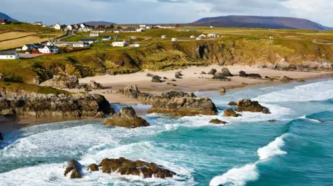 Houses in the village of Durness on the north Highland coast. There are sea cliff down to a beach. The sea is a turquoise colour and waves are crashing in on the shore. 