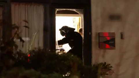 Police officer in black protective suit and helmet stands in a dooraway, lit by orange glow, side-on to camera.