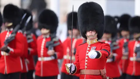 Coldstream Guards can be seen marching with swords and guns, wearing their distinctive red uniform and black bearskin caps, near Buckingham Palace ahead of the procession for the Lying-in State of Queen Elizabeth II on September 14, 2022 in London,