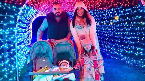 Sophie Jenkins Sophie Jenkins and her husband smiling in a light display, along with Ronnie, one, in a stroller, and Elsie, standing alongside it and in front of her parents