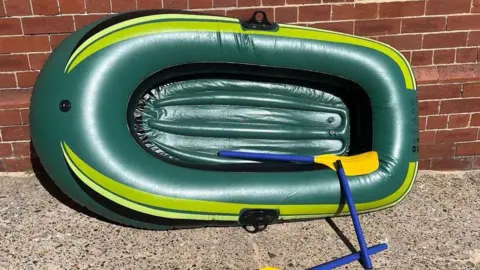 A small inflatable green dinghy with two broken blue oars which has been placed against a brick wall.