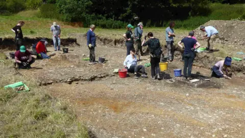 Cerne Historical Society Thirteen people working on an archaeological trench. The remains of a wall can be seen across the centre of the trench