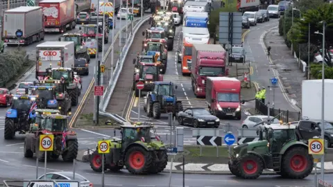 Dozens of tractors driving in Dover. They are going around a roundabout, alongside other vehicles. 