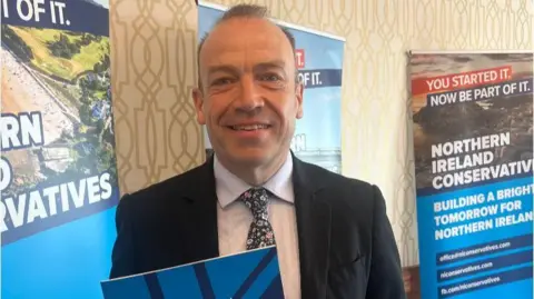 Smiling Chris Heaton-Harris wearing dark jacket, white shirt and patterned tie, holds a copy of a manifesto