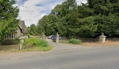 Google Entrance to estate with trees on either side and cars parked near entrance.