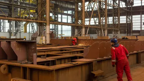 Getty Images A worker grinds pieces of steel during the construction of a barge at the production facility in Harland & Wolff. One workers is wearing a hardhat and the other is wearing a helmet over their eyes. Both are dressed in bright red.