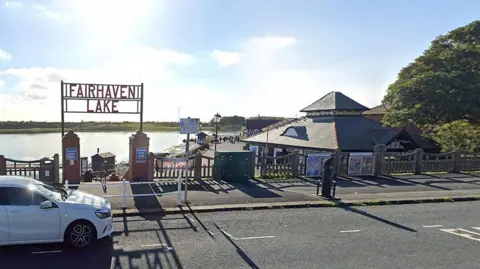 The entrance to Fairhaven Lake. There is a white car parked on a road in the foreground and Fairhaven Lake is in the background.