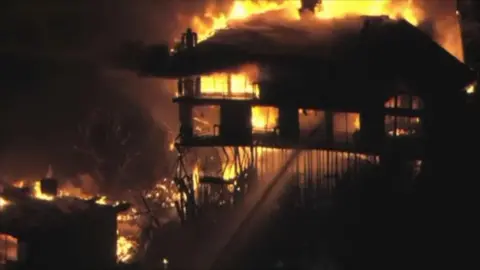 A clip from a video shows water from a fire crew's hose being aimed inside a huge Studio City house which is engulfed in flames at night