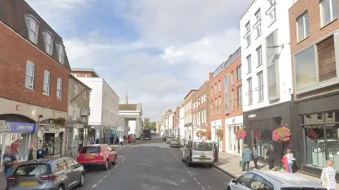 A Google Street view image of a street which is lined by shops on both sides. There are cars parallel-parked next to the pavements.