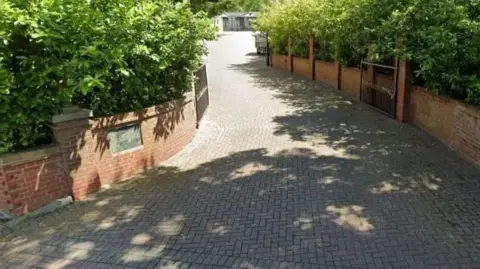 A brick driveway with short brick walls on either side. There are green hedges behind the brick walls