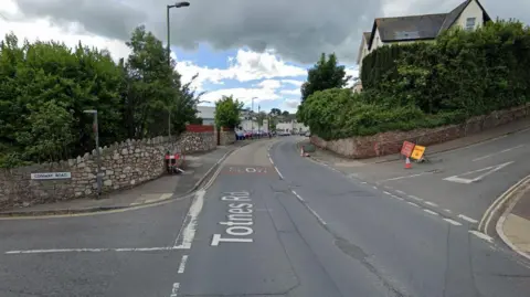 Totnes Road in Paignton at its junctions with Conway Road on the left and Primley Park on the right. A red road closed sign and yellow diversion sign have been set up at the T-junction of Primley Park and are next to a traffic cone. Trees from two gardens are on either side of Totnes Road.