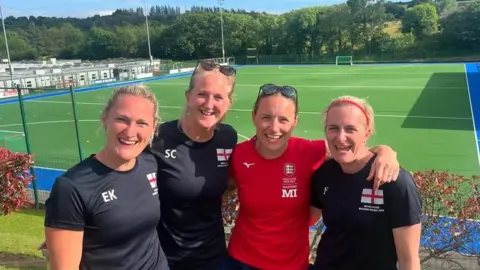 Lucy Field Lucy Field and three of her teammates smile at the camera in front of a hockey pitch. They have their arms around each other and three of them are wearing blue T-shirts and one is wearing a red T-shirt. 