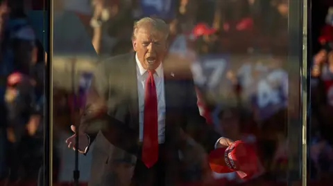 Donald Trump with his hands apart, holding a Make America Great Again cap, on stage and behind bulletproof glass. He is wearing a red tie, white shirt and dark suit, in front of a crowd