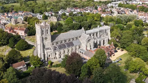 Silversprey An aerial view of Christchurch Priory. The large grey stone building has a large tower at one end with a clock. Behind you can see the ruins of Christchurch Castle and the homes and shops of the town. The area of is full of lush green trees. 