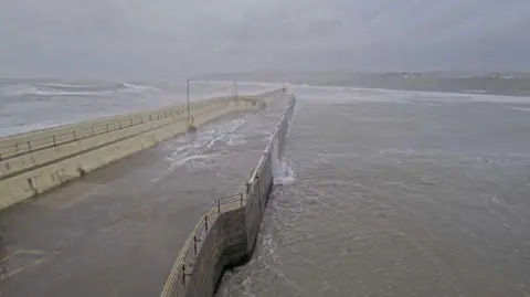 Waves breaking over Peel Breakwater. Water can be seen running back into the sea below in the outer harbour area. The Peel shoreline and Promenade can be seen in the distance.