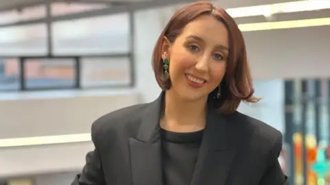 Alex McCormick is smiling and looking at the camera, wearing a grey top and suit jacket. She has red bobbed hair and is wearing green dangly earrings. The library can be seen blurred out behind her.