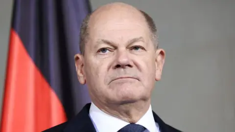 A headshot of Germany's Chancellor Olaf Scholz as he appears expressionless photographed in front of a red and black flag