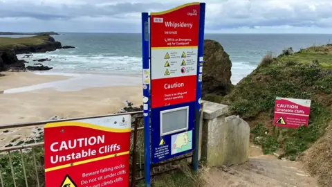 Three signs stand on the top of a cliff with Whipsiderry beach behind and below them. The red warning signs detail the dangers of unstable cliffs and rock falls and have emergency contact details for use in case of an accident.