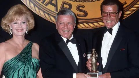 Getty Images Sir Roger smiles as he poses for a picture with singer Frank Sinatra and his wife Barbara. Sir Roger and Frank Sinatra are holding a trophy together. They are dressed smartly in dinner jackets and bow ties. Sir Roger is wearing glasses and looks overjoyed. Frank Sinatra's wife Barbara is standing next to her husband. She has blonde hair, and is wearing a green, off-the-shoulder, evening dress with black tiger stripes on it. She has dangly emerald earrings.