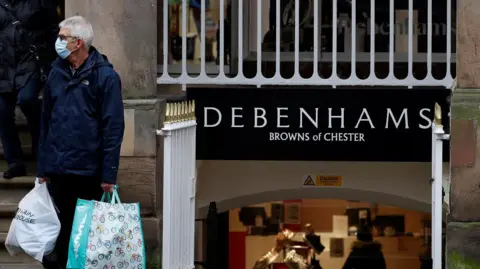 Man wearing a face protection mask and carrying two large shopping bags stands outside the entrance to Debenhams at Browns of Chester