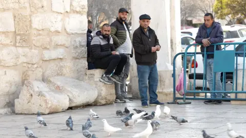 Five men stand or sit by the Nativity Church in Bethlehem, surrounded by pigeons