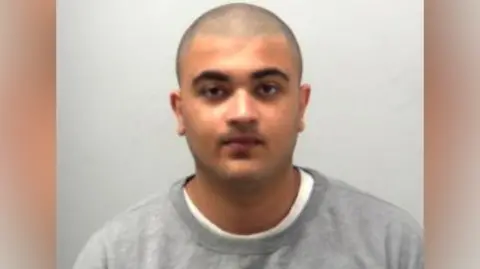 Custody mugshot of Adam Valli. He is wearing a grey sweater with a white T-shirt underneath. He has a shaved head and is expressionless while looking at the camera.