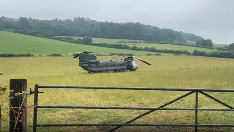 The dark green Chinook with covers on its nose and engine areas in a golden field with a metal gate infront and rolling green fields behind