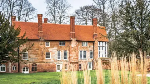 An old-looking large red brick house with tall chimneys with a green garden and tall grasses in the foreground