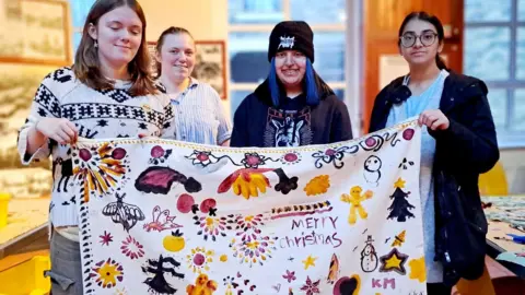 Bradford Council A group of four young women hold up a piece of cloth with yellow, black and pink drawings on it. It also says Merry Christmas.