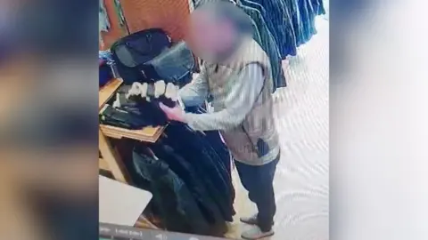 A shoplifter with face blurred gathers up a bundle of green gloves with furry lining from a shelf in a clothes shop.