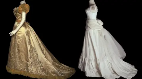 A headless mannequin wears a golden ball gown, with bodice and full skirt, of the late Victorian period, while another wears white undergarments in the form of a corset and skirt.