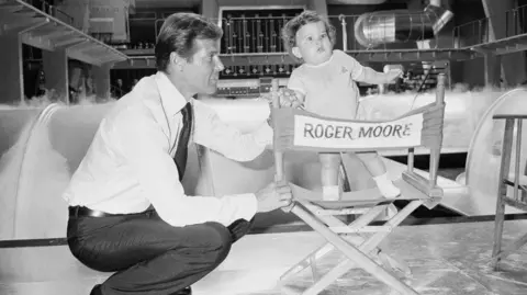 Getty Images In this black and white image, Sir Roger is seen on the film set of the Bond film, The Man With The Golden Gun. He is crouching down next to a chair which has his name written on the back. His young son Christian is standing on the chair and Sir Roger is holding his hand. In the background, you can see pipes and tunnels.