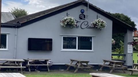 The exterior of The Club at Tuffley Park, which is a grey building with picnic benches outside.